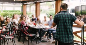 Man standing at a microphone and speaking while patrons enjoy a meal at a restaurant.
